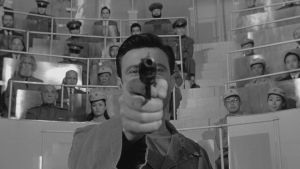 Raymond Shaw (Laurence Harvey) osoittaa pistoolilla suoraan kameraan elokuvassa Mantshurian kandidaatti (1962). Taustalla kiinalaisia ja neuvostoliittolaisia tarkkailijoita