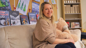 Marianne Falck-Hvilstafeldt. Bilden är tagen i november. En kvinna i beige tröja sitteri en beige soffa bredvid en stor gosebjörn. Bakom henne syns teckningar på julgranar.