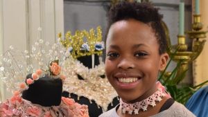 Nosipho Bahne on tehnyt choker-kaulakorun käsin virkatusta pitsistä. Ett porträtt av en leende flicka som har på sig ett spänt halsband, så kallad choker. Halsbandet har hon gjort själv av en rosa spets och sidenband och det är dekorerat med vita, stjärnformade pärlor.