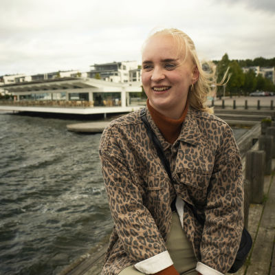 Jasmin Tuomi istuu hymyillen. Taustalla näkyy järven aaltoja ja rakennuksia.