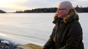 Då Camilla drabbades av allvarlig bröstcancer valde hon att än en gång öppet dela med sig av det svåra i livet. Kvinna sitter på en bänk vid en frusen fjärd. Den låga solen lyser på hennes ansikte. Hon har inget hår på huvudet.