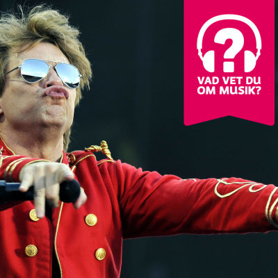 Jon Bon Jovi iklädd solglasögon. Han putar med läpparna, har händerna utsträckta framåt med en mikrofon i höger hand.
