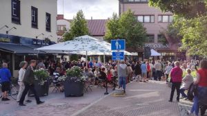 Bild från fjolåret. Mycket människor på en gågatan, de står eller sitter på stolar och bänker. Sommar. Byggnader runt omkring.