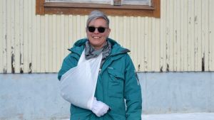 Sirpa Kallio förstår så gott som all svenska, det vill säga standardsvenska. När det kommer till dialekter kan hon snappa upp något ord här och där, men ofta förstår hon väldigt lite av det som sägs. Porträttfoto av kvinna med grönblå jacka, svarta solglasögon, ena armen i mitella. Hon tittar in i kameran och ler stort. Bakom en träbyggnad som är beigemålad.