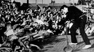 Elvis Presley år 1956. Elvis Presley på scenen år 1956 inför en skrikande pubilk som sträcker ut armarna mot honom.