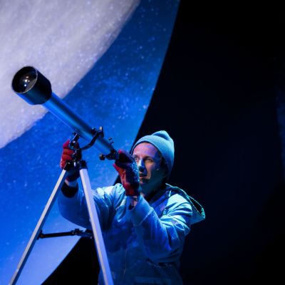 En man i mössa och vantar tittar i en stjärnkikare. I bakgrunden syns en väldig måne. 