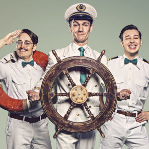 Tre käcka män i sjömansuniformer står på rad bakom ett skeppsroder. 