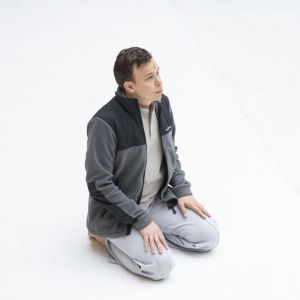 Ruskeatukkainen mies (tanssija Carl Knif) harmaissa verkkareissa ja fleecepuserossa istuu polvillaan lattialla vasten täysin valkoista taustaa ja katsoo yläviistoon oikealle.