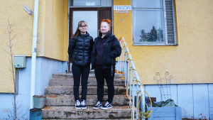 Elin Isaksson och Isabel Virtanen har klätt sig i svart till minne av sin skola som snart inte finns mer. Samtidigt ser de fram emot Prakticums nya fina utrymmen som de ska flytta till. Två unga kvinnor i svarta ytterkläder står på en trappa utanför ett gult hus. På väggen finns en skylt där det står frisör.