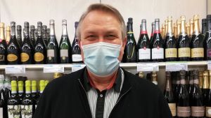 Seppo Metsola, kundservicechef på Alko i Vasa centrum, tackar kunderna för att de underlättar personalens arbete under coronatiden. Mansperson med munskydd ser in i kameran. I bakgrunden vinflaskor på rad i butikshylla.