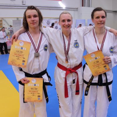 Tre personer i vita taekwondodräkter, medaljer hänger runt halsen och de har diplom i handen.