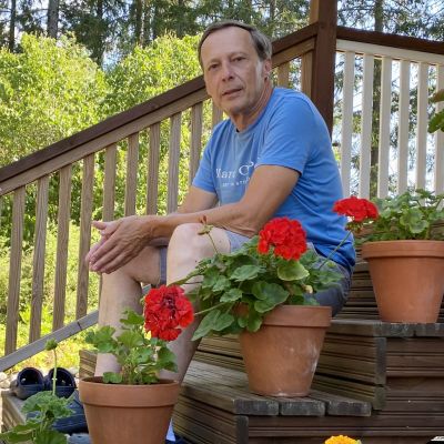En medelålders man i shorts och t-skjorta sitter på en trappa. Bilden är tagen från sidan och nerifrån så att blommorna på trappan syns i förgrunden.