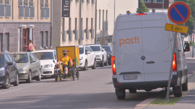 Ett cykelbud med gul skjorta sitter i sin cykel med pedalerna framför sig och en stor gul låda bakom sig. Han cyklar förbi en av Postens bilar.