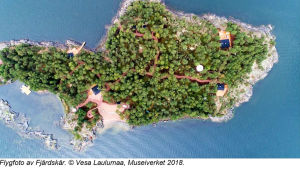 På ett flygfoto från ön syns också den nyanlagda sandstranden nere på bilden. Fotot är inte i nord-sydlig riktning utan norr är till höger. Fotot ingår i planläggningsmaterialet. En flygbild av en ö, Det syns skog, sandstrand, stugor, hav. Solen skiner.