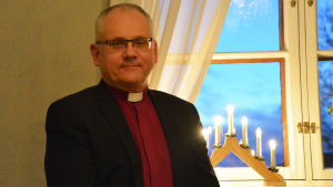 Åstrand tycker inte att man ska sätta ribban alltför högt när det gäller julförberedelserna. Man behöver inte vara perfekt för att få ta del av kärleken och värmen. En medelålders man står invid ett fönster där en elljusstake lyser. Mannen har på sig en svart kavaj, en röd skjorta och en prästkrage.