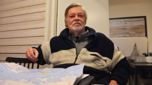 Hasse Hägg har studerat kartan över Sibbo skärgård. En äldre man i randig tröja sitter tillbakalutad på en stol. På bordet framför honom ligger en karta.