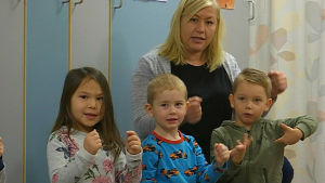 Sandra träffar mellan 40 och 150 barn per dag. Hon undervisar på både daghem, förskolor och skolor i södra Finland. Den här dagen har hon hälsat på hos daghemmet Miili i Sibbo. Tre barn sitter på en bänk och visar rörelser till låten.
