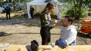 Jimmie Åkesson blir sminkad. Sverigedemokraternas ordförande Jimmie Åkesson blir sminkad vid ett träbord i solskenet, med ett vitt tält i bakgrunden.
