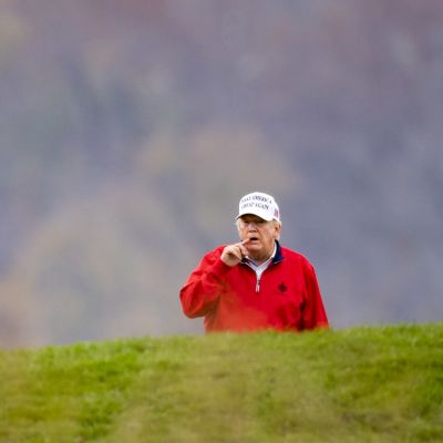 Golfia pelaava Donald Trump katselee väylää kumpareen takaa, kumpareen huipulla on USa:n lipun mallinen golf-lippu. 
