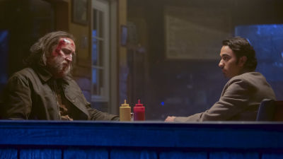 En blodig Rob (Nicolas Cage) och Amir (Alex Wolff) sitter vid ett cafébord och ser allvarliga ut.