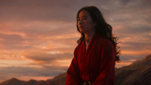 Närbild på Mulan (Yifei Liu) sitter på ett berg i solnedgången.