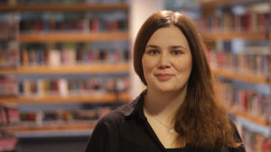 Tanja Fagerholm. En vit kvinna med brunt, långt hår, står framför bokhyllor i ett bibliotek.