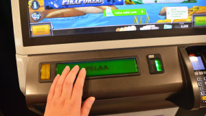 Pelaa, spela, uppmanar automaten. Närbild på en spelautomat och en hand som trycker på en stor knapp där det står pelaa.