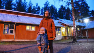 Både mor och son är besvikna över att Kuggom daghem kan komma att stängas. Linda Varoma bor i centrum men har valt att sätta sin son i ett mindre daghem lite utanför centrum. En kvinna i röd jacka står mitt i bild och tittar in i kameran. Hon håller handen på en liten pojkes axel. Han tittar åt vänster, ut ur bilden. Det är mörkt ute. De står framför ett rött trähus. Genom husets glasdörrar syns gult ljus.