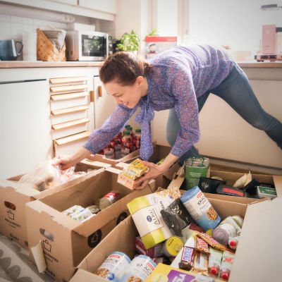En kvinna ler medan hon böjer sig ner och plockar matvaror från lådor i kartong. 