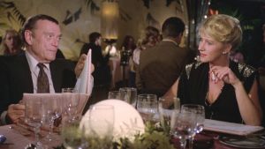 Victoria (Helen Mirren) emännöi eleganttina Atlantin takaa saapunutta Charlieta (Eddie Constantine) Charlie (Eddie Constantine) ja Victoria (Helen Mirren) ravintolan pöydässä elokuvassa Pitkä pitkäperjantai