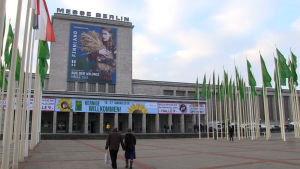 Grüne Woche lockar 400 000 besökare varje år. Två människor vandrar mot en grå byggnad som har skylten Messe Berlin på taket.