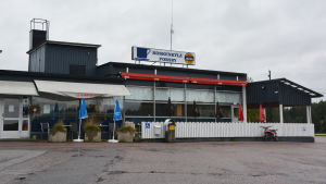 Forsby Café. Ett svart hus med ett vitt lågt staket utanför. Framför huset syns asfalt och bakom huset grå himmel. På taket finns en skylt där det står Koskenkylä Forsby med en bild av en kaffekopp.