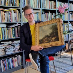 Författaren Peter Mickwitz sittandes framför en bokhylla med en gammal oljemålning i famnen