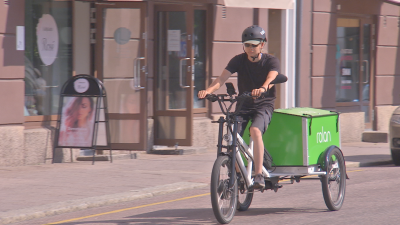 Ett cykelbud med svart hjälm kommer cyklande med en stor grön transportlåda bakpå cykeln.