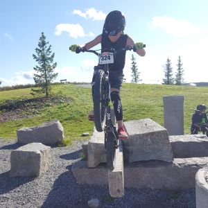 Sisu Kymäläinen går i faderns och farfars cykelspår. Pojke cyklar på en sten vid en grusplan.