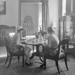 Smått, nätt och stämningsfullt vid bordet. Café i Helsingfors på 1930-talet. Svartvit bild på två kvinnor som sitter vid ett litet runt kaffebord på ett café.