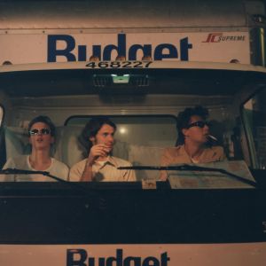 Kolme nuorta miestä istuu vierekkäin muuttoauton etupenkillä. Kuva on otettu etulasin lävitse. Kuljettaja polttaa tupakkaa tummissa aurinkolaseissa. Konepellissä lukee "Budget".