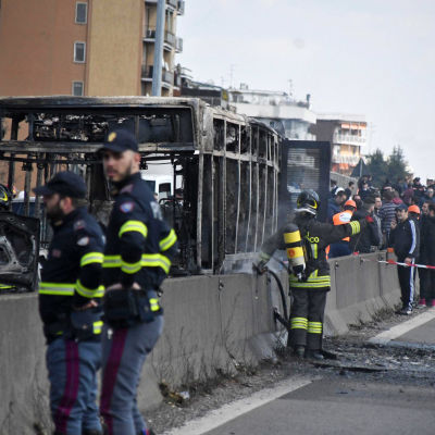 brandmän står framför en uppbränd buss. Två i bildens förgrund och en i bakgrunden.