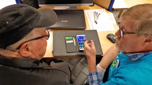 Mies opastaa toista tietotekniikassa Kajaanin kirjastolla Nettiä ikä kaikki -tapahtumassa. Pöydällä kännykkä auki.