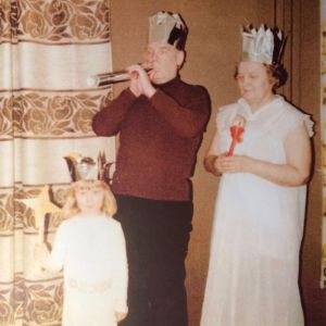 Pikkutyttö ja isovanhemmat yhteisessä esiintymisleikissä, kaikilla kultapahviset kruunut päässä.