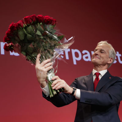 En man i grått hår håller i en bukett med röda rosor och ler.
