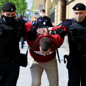 En man grips av polis. Han står mellan två poliser, nerböjd medan de håller ner honom.