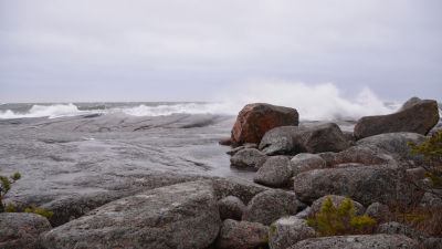 En strand med stora släta stenar. Stora vågor slår mot stenarna och himlen är grå.