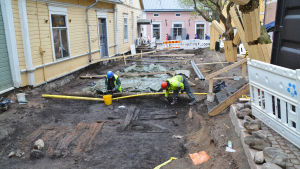 Hela halva gränden har grävts upp Arkeologerna har än så länge bara hunnit undersöka en del av området närmast kameran. En gränd i Gamla stan i Borgå har grävts upp. Två personer i hjälmar och gula västen gräver i den stora gropen mellan husen. Stora rör går tvärs över gropen.