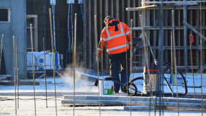 Byggarbetare är en vanlig syn i Majberget under vardagar. En byggarbetare i röd jacka jobbar på en byggarbetsplats. Han står med ryggen mot kameran och ser ut att hålla i en borste. Bakom honom finns en målfärgsburk och en dammsugare.