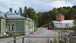 Det gamla stationshuset i grönt står strax intill lokstallet. Till vänster syns ett stort grönt hus, den gamla järnvägsstationen. På andra sidan vägen, till höger, finns ett tegelhus med ett torn. Taket är intäckt med vita presenningar.