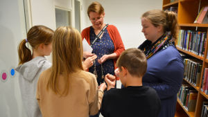 Heli Salminen och Bettina Bergström fäster spelarna vid varandra med leksakshandklovar. Det kräver bra samarbete för att utforska rummet medan spelarna hänger ihop. Tre barn står framför två vuxna kvinnor. Kvinnorna håller på med att sätta handklovar på barnen.