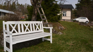 Här kunde det vara skönt att sitta. I förgrunden synns en vit träbänk i en trägård. Bakom den ett trähus och en vit bil.