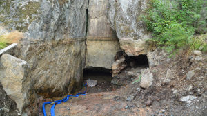 Michel Merle berättar att de har pumpat ut vatten från grottan där Lemminkäinens tempel påstås ligga och att de till följande planerar att fixa till grottan på utsidan så att den blir fin. En grotta med två låga öppningar. En blå slang sträcker sig in i grottan.