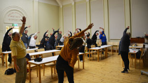 Diskussionsmötet börjades med lite stretchning. Flera personer står upp i ett mötesutrymme och stretchar med armarna i luften.